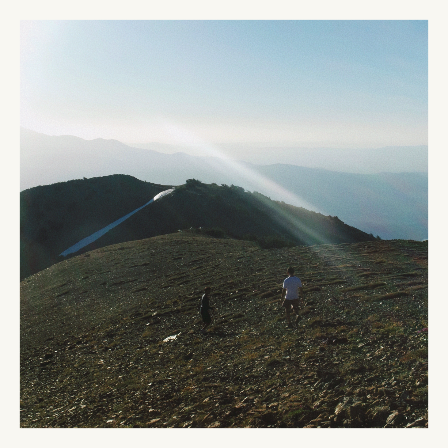 Two people crossing a mountain ridge beneath pale blue sky and sunbeams